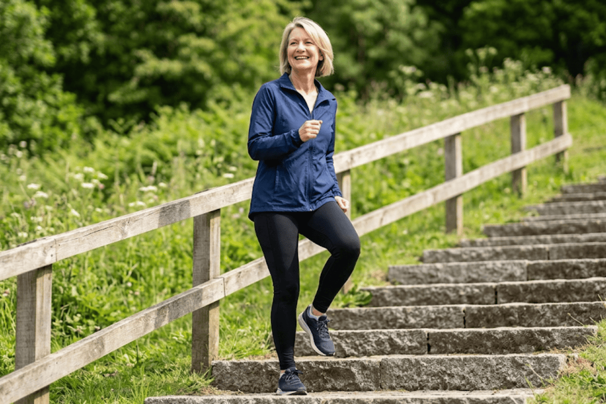 Smiling woman in a blue jacket and black leggings jogging up outdoor stone stairs beside a wooden railing in a green park setting.